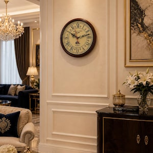 May include: A round wall clock with a dark brown wooden frame and a cream-colored face. The clock features black Roman numerals and decorative hands. The clock is mounted on a light beige wall in a living room setting.