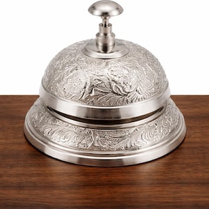 May include: A silver-colored desk bell with an ornate floral pattern. The bell has a rounded top with a small handle and sits on a circular base. The bell is displayed on a dark brown wooden surface.
