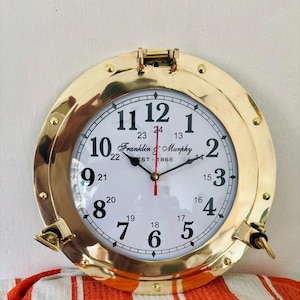 Könnte beinhalten: Eine runde Messing-Uhr mit weißem Zifferblatt und schwarzen Ziffern. Die Uhr hat ein nautisches Bullauge-Design mit einem aufklappbaren Deckel und seitlichen Verschlüssen. Das Zifferblatt zeigt "Franklin & Murphy EST-1868".