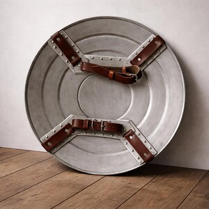 May include: A circular, silver-toned shield featuring brown leather straps and rivets. The shield's design includes a central circle and a cross-shaped support on the reverse. The leather straps are fastened with buckles for a secure grip.