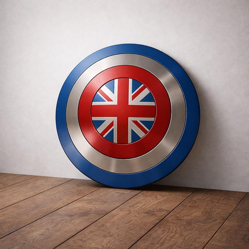 May include: A circular shield featuring the Union Jack flag at its centre, surrounded by a red ring, a silver band, and a blue outer rim. The shield is propped against a white wall on a wooden floor.