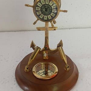 May include: A nautical-themed desk clock and compass set. The clock, shaped like a ship's wheel, features Roman numerals and is mounted on an anchor. A compass is set into a wooden base, with two pen holders. The clock and compass are made of brass.