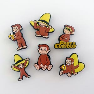 Puede incluir: Una colección de seis dijes para zapatos marrones y amarillos con un mono de dibujos animados en varias poses. Algunos dijes muestran al mono con un sombrero amarillo, gafas de sol o sosteniendo un plátano. Un dije tiene el texto "Fast & Curious".