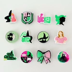May include: A collection of twelve colorful shoe charms featuring designs and text from the musical "Wicked." Charms include images of witches, the show's logo, and other related graphics in shades of green, pink, black, and white. The charms are arranged on a white surface.