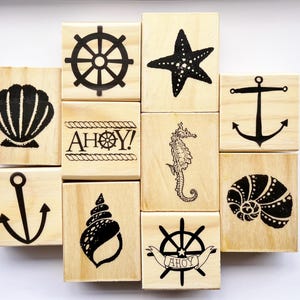 Puede incluir: Bloques de madera con diseños náuticos en negro. Incluyen ilustraciones de conchas, timones, estrellas de mar, anclas, caballitos de mar y el texto "AHOY!".