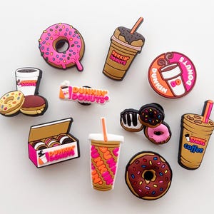 Peut inclure: Une collection de breloques colorées sur le thème de Dunkin' Donuts, comprenant des beignets avec glaçage rose, marron et chocolat, des tasses à café et une boîte de beignets. Les breloques présentent le logo Dunkin' Donuts et divers motifs.