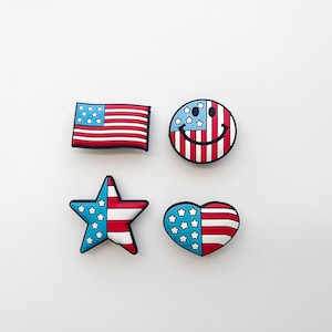 Puede incluir: Cuatro dijes patrióticos en rojo, blanco y azul. El conjunto incluye una bandera americana, una carita sonriente con elementos de la bandera, una estrella y un corazón, todos con un contorno negro. Ideal para añadir un toque de Americana a los accesorios.