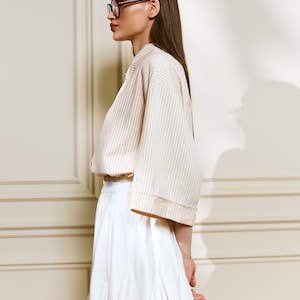 May include: A woman wearing a cream-colored, striped, short-sleeved top and a white pleated skirt. The top has a relaxed fit, and the skirt falls to mid-thigh. She is wearing glasses and has long, dark hair. The outfit is simple and elegant.