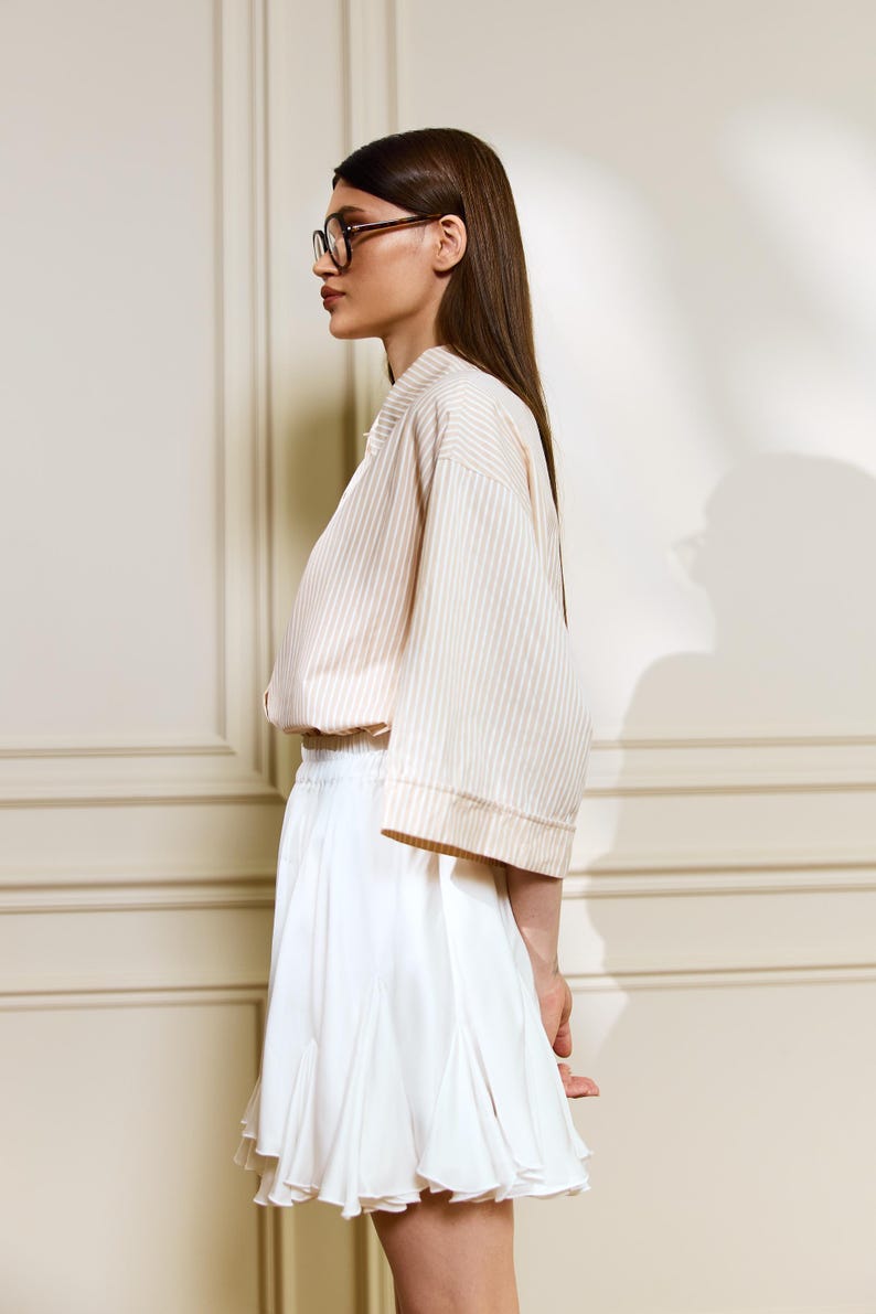 May include: A woman wearing a cream-colored, striped, short-sleeved top and a white pleated skirt. The top has a relaxed fit, and the skirt falls to mid-thigh. She is wearing glasses and has long, dark hair. The outfit is simple and elegant.