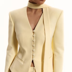 Pode incluir: Um blazer amarelo-p&aacute;lido com decote em V profundo, mangas compridas e fecho frontal com seis bot&otilde;es revestidos a tecido a condizer. A pe&ccedil;a inclui uma gravata comprida integrada tipo cachecol e dois bolsos horizontais na cintura.
