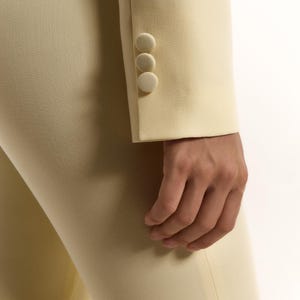 Pode incluir: Uma manga de blazer amarelo-p&aacute;lido com tr&ecirc;s bot&otilde;es a condizer revestidos a tecido, alinhados verticalmente no punho. A manga faz parte de uma pe&ccedil;a de vestu&aacute;rio ajustada feita de tecido liso e s&oacute;lido, com linhas limpas e corte estruturado.