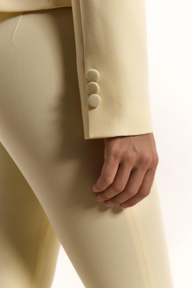 Pode incluir: Uma manga de blazer amarelo-p&aacute;lido com tr&ecirc;s bot&otilde;es a condizer revestidos a tecido, alinhados verticalmente no punho. A manga faz parte de uma pe&ccedil;a de vestu&aacute;rio ajustada feita de tecido liso e s&oacute;lido, com linhas limpas e corte estruturado.
