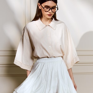 May include: A woman wearing a beige and white striped button-down shirt with a white skirt. The shirt has wide sleeves and a collared neckline. The skirt is short and flared. She is wearing glasses.