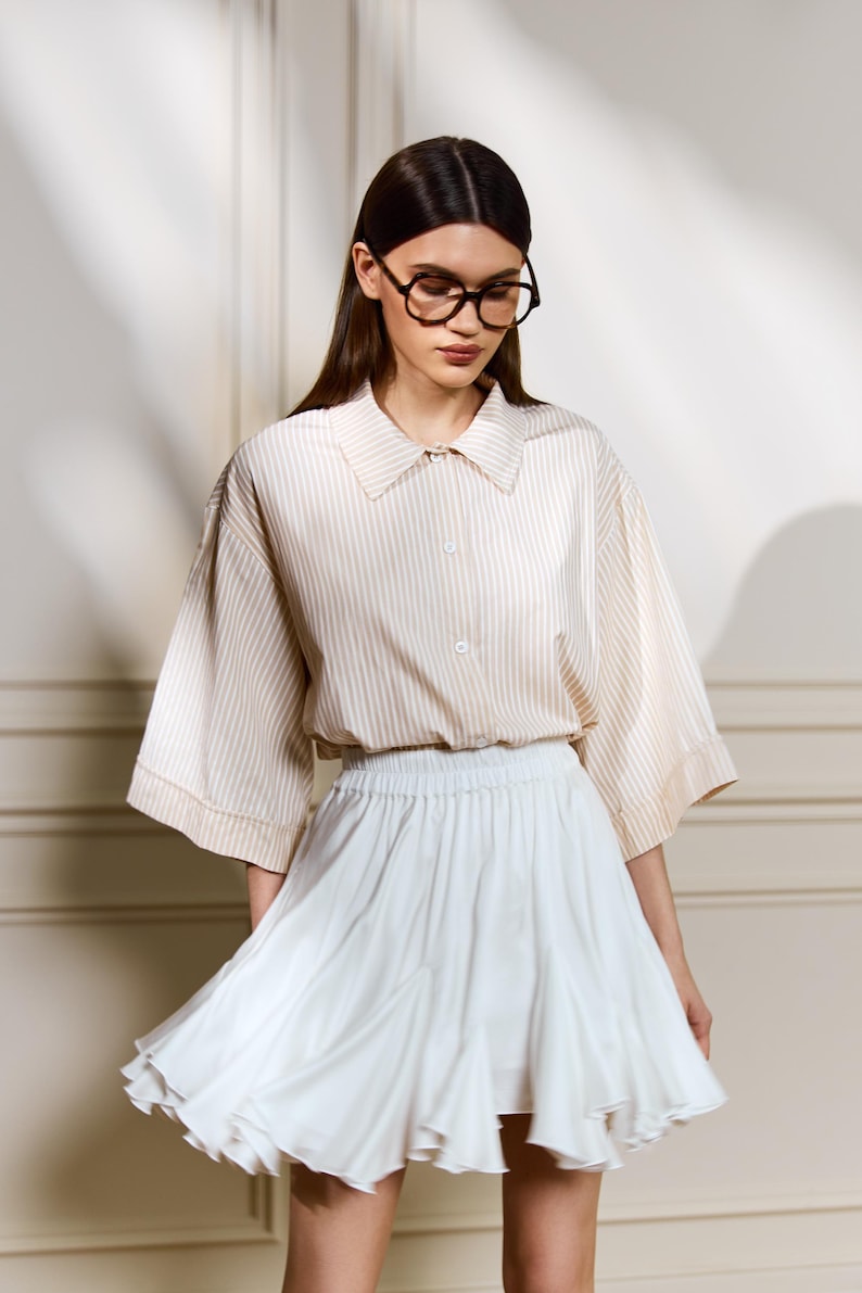 May include: A woman wearing a beige and white striped button-down shirt with a white skirt. The shirt has wide sleeves and a collared neckline. The skirt is short and flared. She is wearing glasses.