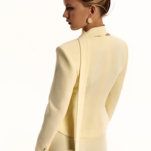 Pode incluir: Um blazer amarelo-p&aacute;lido com gola alta e um longo painel de tecido tipo cachecol que cai pelas costas. O blazer tem mangas compridas com tr&ecirc;s pequenos bot&otilde;es nos punhos e &eacute; combinado com cal&ccedil;as a condizer em amarelo-p&aacute;lido.