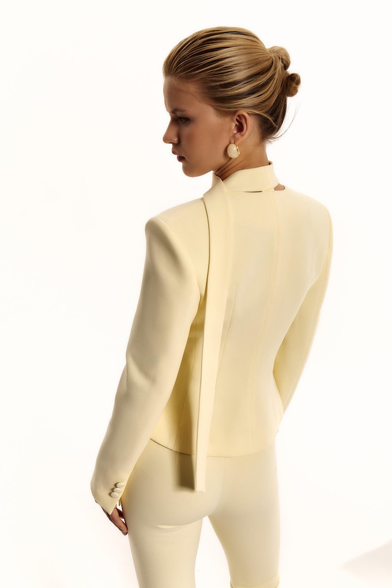 Pode incluir: Um blazer amarelo-p&aacute;lido com gola alta e um longo painel de tecido tipo cachecol que cai pelas costas. O blazer tem mangas compridas com tr&ecirc;s pequenos bot&otilde;es nos punhos e &eacute; combinado com cal&ccedil;as a condizer em amarelo-p&aacute;lido.