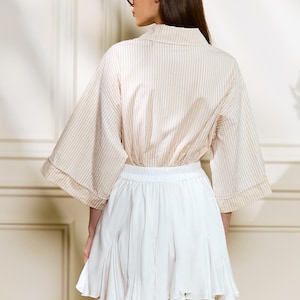 May include: A beige and white striped blouse with wide sleeves and a white pleated skirt. The blouse has a collared neckline. The skirt is short and flared. The outfit is suitable for casual or semi-formal wear.