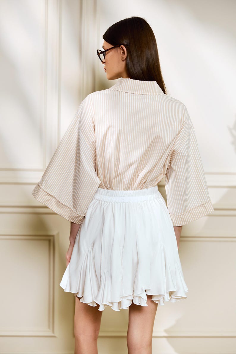 May include: A beige and white striped blouse with wide sleeves and a white pleated skirt. The blouse has a collared neckline. The skirt is short and flared. The outfit is suitable for casual or semi-formal wear.