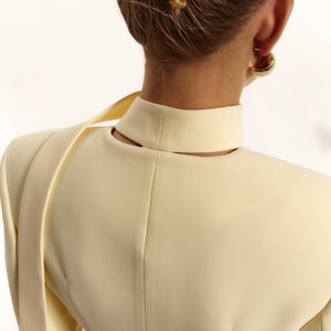 Pode incluir: Um blazer amarelo-p&aacute;lido com gola alta e um detalhe recortado na parte de tr&aacute;s do pesco&ccedil;o. A pe&ccedil;a tem ombros estruturados e uma tira de tecido longa e fina que se estende a partir da zona do ombro, vis&iacute;vel a partir da vista traseira.