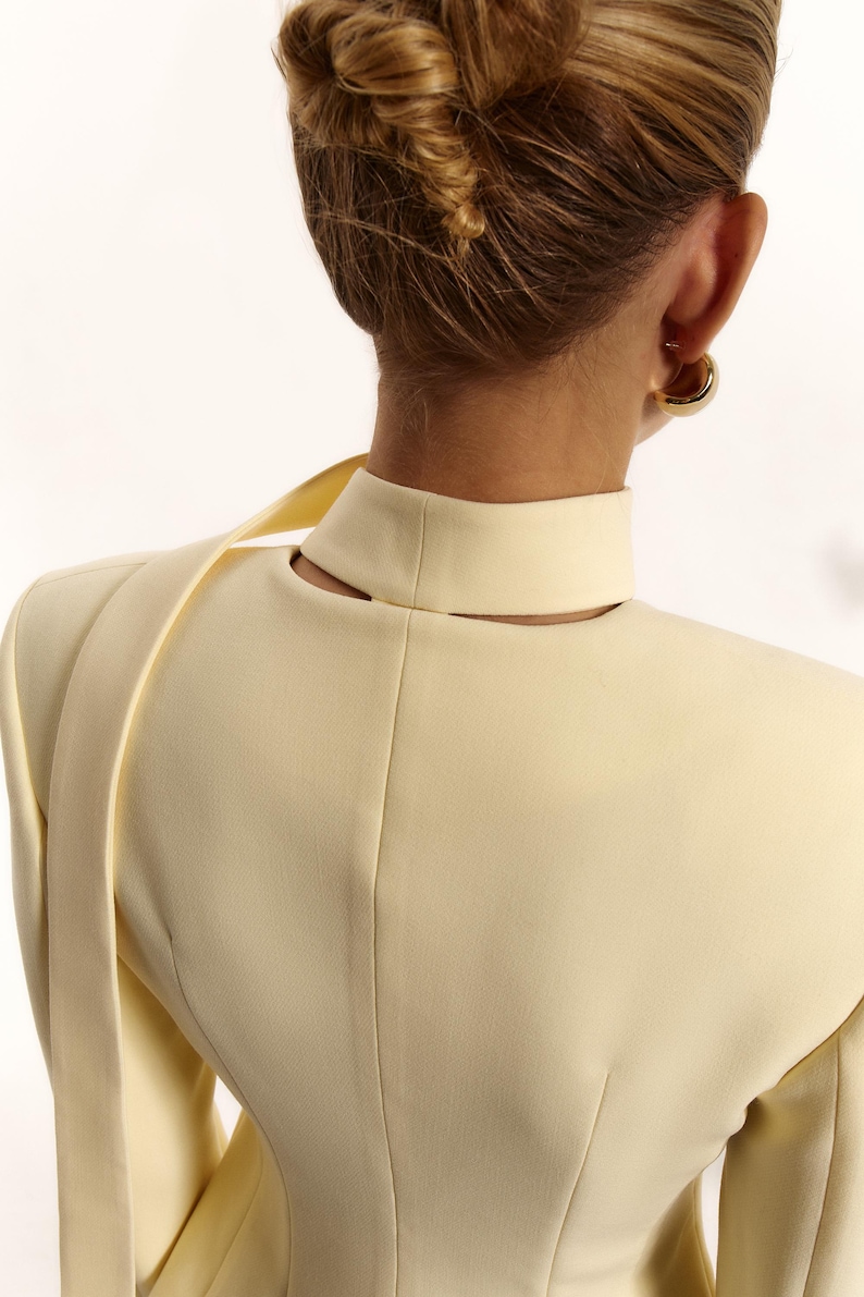 Pode incluir: Um blazer amarelo-p&aacute;lido com gola alta e um detalhe recortado na parte de tr&aacute;s do pesco&ccedil;o. A pe&ccedil;a tem ombros estruturados e uma tira de tecido longa e fina que se estende a partir da zona do ombro, vis&iacute;vel a partir da vista traseira.