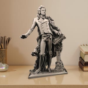 Può includere: Statua color argento di un uomo in abito storico, con un libro in mano e che gesticola con la mano destra. La statua presenta dettagli intricati nei vestiti e nei lineamenti del viso e poggia su una base decorativa.
