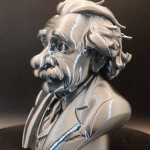 May include: A silver-coloured bust of Albert Einstein, depicting his distinctive hairstyle, moustache, and suit. The sculpture is detailed, with visible lines and textures, against a dark backdrop.
