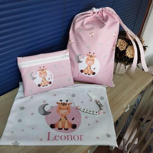 Pode incluir: Um conjunto de acessórios para bebés com um design de girafa. O conjunto inclui um saco rosa com cordão, uma bolsa com zíper e um saco plano, todos adornados com uma girafa de desenho animado, uma lua, estrelas e o nome "Leonor".