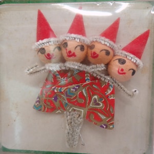 May include: Four small elf ornaments with wooden bead heads, painted facial features, and red felt cone hats with white trim. Each elf has a white pipe cleaner collar and a body made of red patterned paper, all encased in clear plastic packaging.