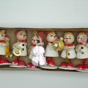 May include: A set of six vintage-style snowman figurines arranged in a cardboard box. Each snowman wears a red hat and scarf, has a round wooden head with painted features, and holds a miniature musical instrument, including a horn, cymbals, cello, drum, and trumpet.