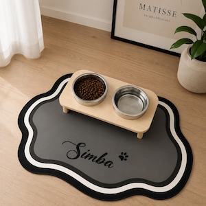 Puede incluir: Un soporte de madera clara para mascotas con dos cuencos de acero inoxidable, uno lleno de pienso marrón, sobre una alfombrilla gris, negra y blanca de bordes festoneados con el nombre Simba y un pequeño gráfico de huella en negro.