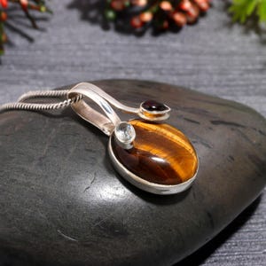 Könnte beinhalten: Silberanhänger mit einem ovalen Tigerauge-Stein, einem kleinen runden roten Stein und einem kleinen runden blauen Stein. Der Anhänger befindet sich an einer Silberkette. Der Tigerauge-Stein ist braun mit goldenen Streifen.