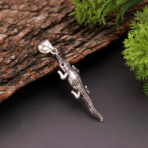Op de afbeelding: Een zilverkleurige hanger in de vorm van een alligator. De hanger heeft een getextureerd lichaam en gedetailleerde kenmerken, waaronder kleine ogen en poten. De alligator hangt aan een kleine lus aan de bovenkant.