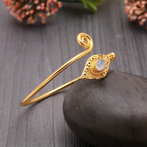 Puede incluir: Un brazalete dorado con un diseño en forma de hoja y una piedra central ovalada de color claro. El brazalete presenta un detalle en espiral en la parte superior y descansa sobre una piedra gris oscura.