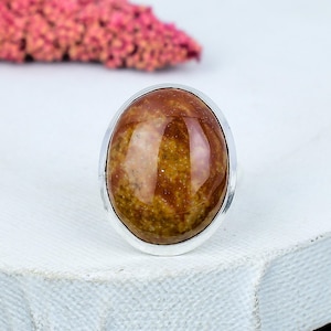 Puede incluir: Anillo de piedra preciosa ovalada con una banda plateada. La piedra preciosa presenta una mezcla de tonos marrones y beige, con una superficie pulida y brillante. El anillo está sobre un fondo blanco, con un elemento floral rosa borroso en el fondo.