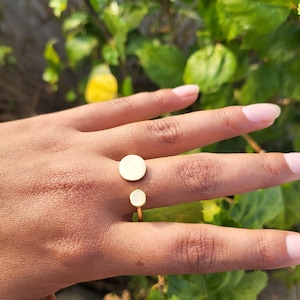 Può includere: Un anello color oro con due elementi circolari bianco sporco. L'anello presenta un design minimalista con un piccolo cerchio sotto un cerchio più grande, collegati da una sottile fascia. L'anello è mostrato su una mano con uno sfondo naturale.