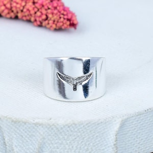 Può includere: Un anello largo e argentato con un design a coda di balena. La coda è rifinita con piccoli abbellimenti scintillanti. L'anello ha una finitura liscia e lucida e un'estetica minimalista. L'anello è un gioiello.