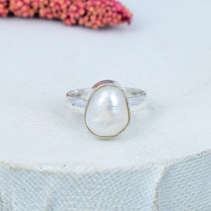 May include: A silver ring with a large, white, irregularly shaped pearl. The pearl is set in a silver band, creating a classic design. The ring is displayed on a white surface, with a blurred pink floral element in the background.