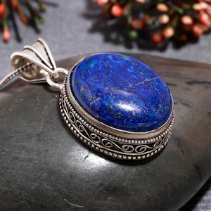 Puede incluir: Collar con colgante de plata con una piedra preciosa ovalada de color azul oscuro con motas doradas. La piedra preciosa está engastada en un bisel de plata decorativo con un diseño calado ornamentado. El colgante cuelga de una delicada cadena de plata.