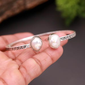 Pode incluir: Uma pulseira de punho prateada com duas grandes pérolas brancas de formato irregular. A pulseira tem um design delicado e padronizado ao longo das bordas. As pérolas são fixadas em uma moldura de bisel, adicionando um toque de elegância a esta joia.