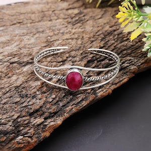 Può includere: Un bracciale a polsino argentato con una grande gemma rossa ovale al centro. Il bracciale presenta un design intrecciato strutturato sui lati e una finitura liscia e lucida. Il gioiello è esposto su una superficie di legno rustico.