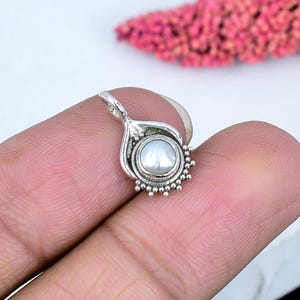 Può includere: Un piccolo ciondolo in argento con una perla bianca rotonda al centro. Il ciondolo ha un design decorativo di ispirazione floreale con piccoli accenti di perline attorno alla perla. L'anello è un anello per una catena.