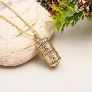Könnte beinhalten: Eine goldfarbene Halskette mit Anhänger, die einen zylindrischen Anhänger mit aufwendigen geprägten Mustern aufweist. Der Anhänger hängt an einer filigranen Goldkette. Der Anhänger hat ein detailliertes Design mit floralen und geometrischen Elementen.