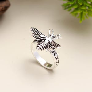 Può includere: Anello a forma di ape argentato. L'anello presenta un design dettagliato a forma di ape con ali e corpo strutturati. La fascia ha un motivo decorativo. L'anello è posto su uno sfondo neutro, evidenziando i dettagli del gioiello.