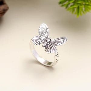 Può includere: Un anello color argento con un design a farfalla dettagliato. La farfalla ha ali strutturate ed è il punto focale dell'anello. La fascia ha un motivo strutturato. Questo è un gioiello.
