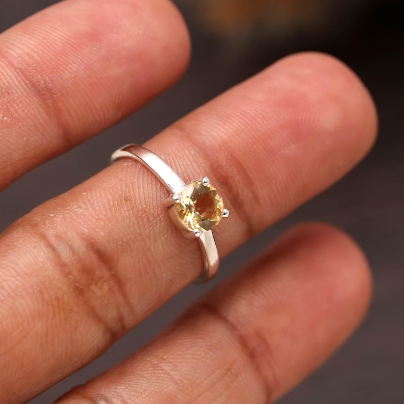 Puede incluir: Un anillo de plata con una piedra preciosa de citrino amarillo redonda y facetada, engastada en una montura de cuatro garras. La banda es lisa y pulida, mostrando un dise&ntilde;o sencillo que resalta la claridad y el tono dorado de la piedra.