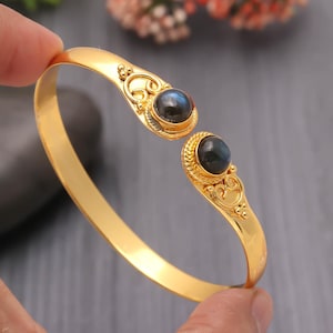 Könnte beinhalten: Ein goldfarbenes Armreif-Armband mit zwei dunklen, runden Edelsteinen. Das Armband hat ein offenes Design mit aufwendigen Details um die Edelsteine. Die Edelsteine sind in einer dekorativen, strukturierten Fassung gefasst.