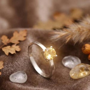 Puede incluir: Un anillo de plata con una piedra preciosa de color amarillo p&aacute;lido, redonda y facetada, montada en un engaste de cuatro garras. El aro es liso y met&aacute;lico, con un acabado pulido que refleja la luz. La piedra es clara y circular.