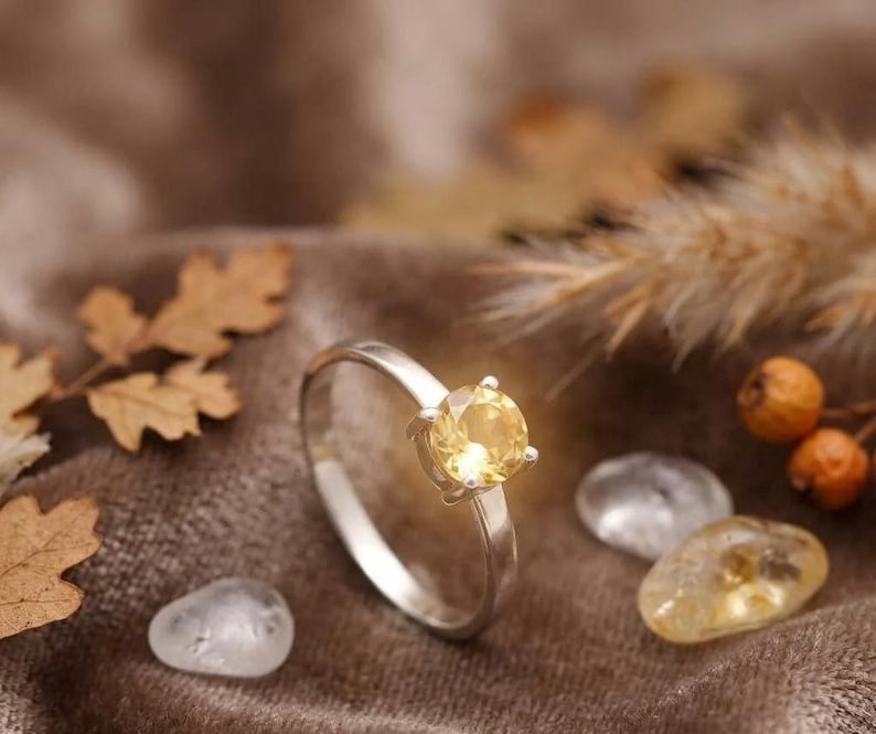 Puede incluir: Un anillo de plata con una piedra preciosa de color amarillo p&aacute;lido, redonda y facetada, montada en un engaste de cuatro garras. El aro es liso y met&aacute;lico, con un acabado pulido que refleja la luz. La piedra es clara y circular.