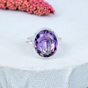 Può includere: Un anello d'argento con una grande ametista ovale. L'ametista è di colore viola intenso e ha una superficie sfaccettata. L'anello è posto su una superficie bianca, con un elemento floreale rosa sfocato sullo sfondo.