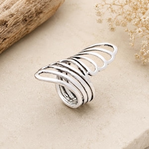 May include: A silver-toned metal ring featuring an open, wrap-around design with multiple curved, parallel bands that extend outward to form a fan-like shape. The surface of the metal has a slightly oxidized, textured finish with dark accents.